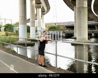 Giovane uomo accanto al fiume, poggiando su ringhiere, utilizzando un lettore MP3 collegato al braccio Foto Stock