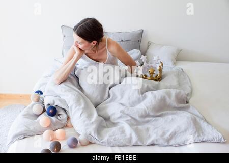 Donna matura nel letto sotto la trapunta con luci decorative mano sulla bocca che guarda lontano Foto Stock