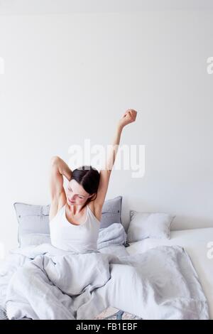 Donna matura indossando vest seduta nel letto sotto la trapunta a braccia alzate stretching Foto Stock