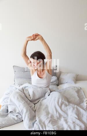 Donna matura indossando vest seduta nel letto sotto la trapunta a braccia alzate stretching Foto Stock