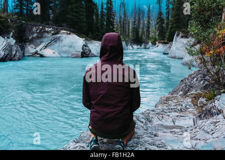 Vista posteriore metà uomo adulto che indossa cappuccio impermeabile accovacciato waters edge Moraine Lake il Parco Nazionale di Banff Alberta Canada Foto Stock