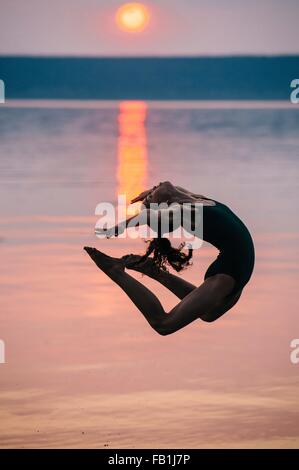 Vista laterale della ragazza dall' oceano al tramonto, saltando a metà in aria flessione all'indietro Foto Stock