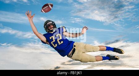 Un adolescente di sesso maschile giocatore di football americano raggiungendo per la cattura di sfera metà aria contro il cielo blu Foto Stock