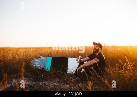 Surfista maschio a bere birra nel campo di erba lunga al tramonto, San Luis Obispo, California, Stati Uniti d'America Foto Stock