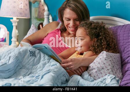 Madre e figlia coccole a letto libro lettura sorridente Foto Stock
