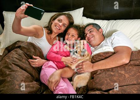 Angolo di Alta Vista della ragazza nel letto con i genitori e il cane utilizza lo smartphone per prendere selfie sorridente Foto Stock