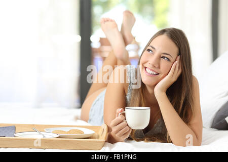 Felice ragazza avente la colazione tenendo una tazza di caffè giacente sul letto e guardando lateralmente Foto Stock