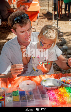 In corrispondenza di un dehor estivo festival in Rancho Santa Margarita, CA, a 5 anno vecchia ragazza stringhe gioielli perline con l aiuto di suo padre. Nota face painting e la selezione di perle in primo piano. Foto Stock