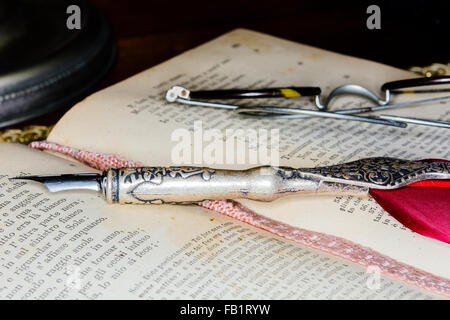 Antica penna stilografica su un vecchio libro e bicchieri. Sfondo scuro in ombra. Foto Stock