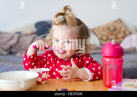 18 mese Baby girl mangiare yogurt da se stessa con un cucchiaio Foto Stock