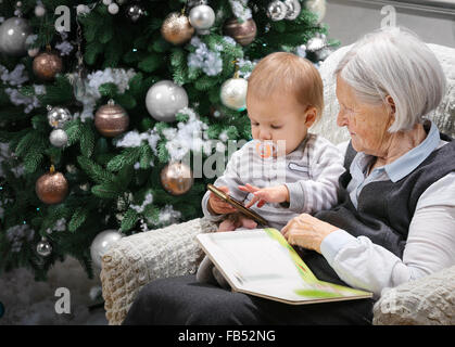 Senior donna la lettura di un libro di suo nipote bambino accanto a un albero di Natale, il ragazzo sta giocando con uno smartphone Foto Stock