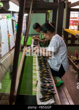 Due donne tessitura utilizzando navette con bobine di filo su di un telaio di tessitura. Parzialmente in tessuto di seta verde visibile del tessuto. Mandalay, Myanmar. Foto Stock