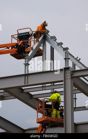 I lavori di costruzione nel Regno Unito : uomini al lavoro in altezza su elevato 'cherry picker' piattaforme, imbullonando insieme il telaio di acciaio di un nuovo sviluppo per il supermercato Tesco e Marks & Spencer store su un sito di costruzione in Aberystwyth, Wales UK Foto Stock