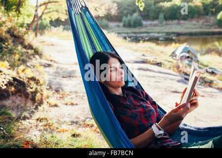 Giovane donna libro lettura sdraiati sulle amache in foresta Foto Stock