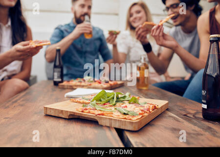 Immagine ravvicinata di pizza sul piatto di legno con persone di mangiare e di bere in background. Gruppo di amici riuniti attorno al tavolo Foto Stock