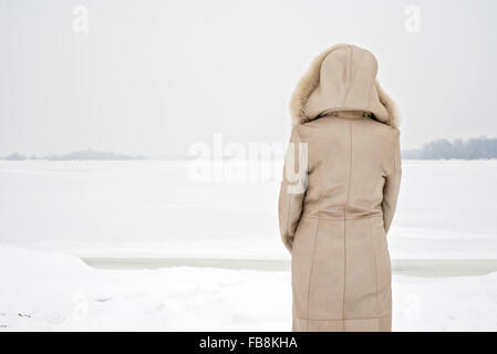 Una donna con un cappotto di pelle con una cappa di pelliccia sta guardando la congelati fiume Dnieper durante un freddo e grigio triste inverno mattina und Foto Stock