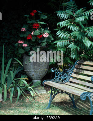 Rosa e rosso dei gerani in un urna di pietra accanto a una vecchia panchina da giardino in un tranquillo angolo del giardino Foto Stock