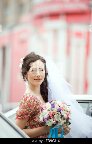 La sposa in semplice abito retrò con motivo floreale, già di indossare il velo ed azienda bouquet nuziali, fiori viola, pone al di fuori della vettura, telaio verticale. Guardando dritto in telecamera. Foto Stock