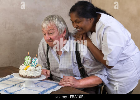 Uomo anziano in casa di cura soffiando le candeline sulla sua torta di compleanno Foto Stock