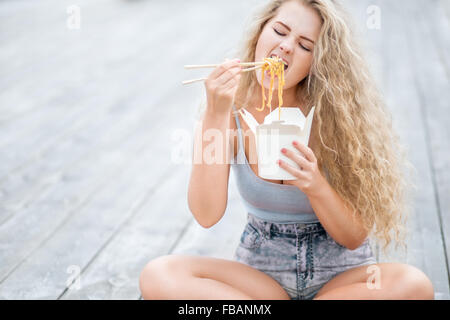 Felice giovane donna con lunghi capelli ricci, tenendo una scatola di pranzo e mangiare tagliatelle cinesi take-out con bacchette. Foto Stock