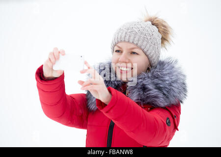 Felice bella ragazza in berretto lavorato a maglia e rosso cappotto invernale utilizzando il telefono cellulare e di scattare una foto con lo smartphone all'aperto contro il Foto Stock