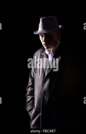 Ritratto del misterioso uomo caucasico in black hat e ricoprire con espressione dura camminare nelle tenebre, chiave di basso studio shot contro Foto Stock