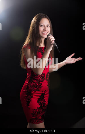 Ritratto di sorridente bella giovane femmina altoparlante con microfono in abito rosso in piedi in Spotlight Foto Stock