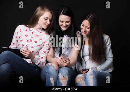 Gruppo di bella ragazza seduta con diversi dispositivi, cercando insieme a uno dei telefoni cellulari e ridere, migliori f Foto Stock