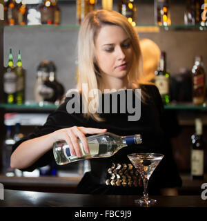 Bella bionda barista ragazza che serve drink alcolici al banco bar, versando bevande in bicchiere da cocktail, concentrarsi sulla bottiglia in mano Foto Stock