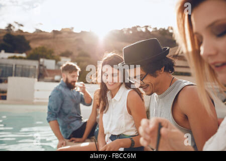 Colpo all'aperto di felice giovane uomo seduto a bordo piscina con i suoi amici di festa. Un gruppo di giovani appeso attorno a una piscina p Foto Stock
