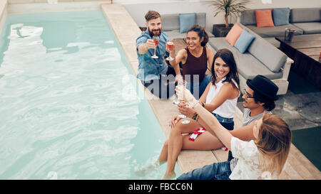 Alta angolazione dei giovani seduti a bordo piscina con vino e sorridente. Gruppo di amici e multirazziale la tostatura in party in piscina Foto Stock