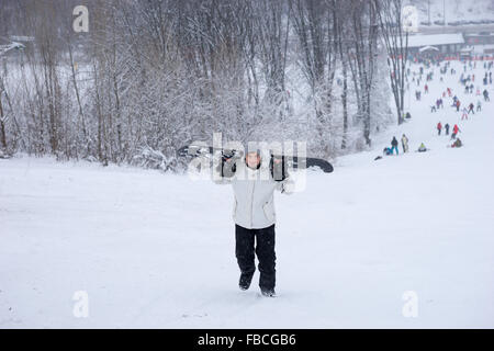 Giovane uomo trascinarsi faticosamente attraverso la neve con il suo snowboard a scalare una montagna verso la telecamera con una folla di persone visibili sulla neve eseguire nella distanza. Foto Stock