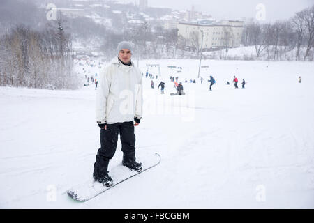 Giovane uomo in piedi sul suo snowboard su una pista da sci guardando la telecamera come egli si prepara a fare una discesa con gli sciatori visibile dietro. Foto Stock