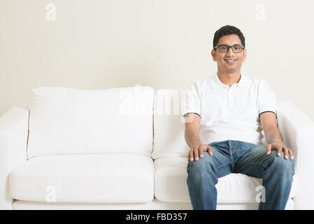 Giovane singolo ragazzo indiano seduto solitario sul divano e sorridente. Stile di vita uomo asiatico a casa. Bel modello maschile. Foto Stock