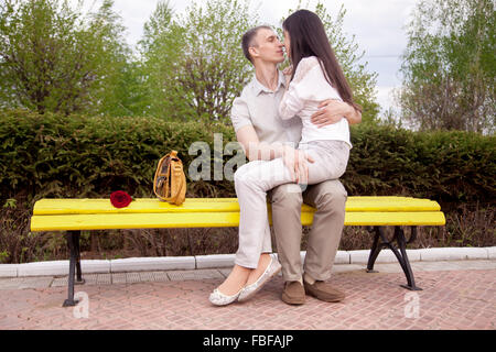 Ritratto di giovane con una rosa rossa baciare sulla panchina giallo nel parco, giovane donna seduta sul suo fidanzato giri a piena lunghezza Foto Stock