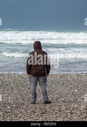 Uomo in piedi su una spiaggia di ciottoli a riva guardando il mare Foto Stock