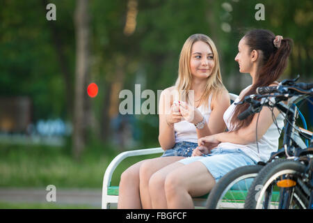 Due sorridenti bella giovane biker donne amici indossando abiti casual seduto su una panchina nel parco il giorno di estate, chat, provvista di Foto Stock