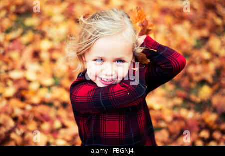 Carino dai capelli biondi ragazza con occhi blu e rosso e nero maglietta controllati circa a gettare le foglie d'arancio in autunno Foto Stock