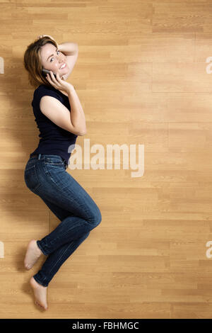 Donna sorridente giacente sul suo lato sinistro sul pavimento, appoggiata sul suo gomito, con il suo cellulare al suo orecchio destro. Foto Stock