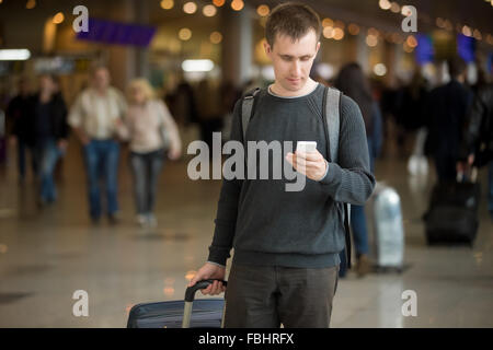 Ritratto di giovane uomo bello in 20s a piedi in aeroporto moderno terminale tramite smart phone app in pubblico wifi area, sms, tr Foto Stock