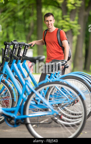 Giovani sorridenti man standing a noleggiabile city bike parking lot, scegliendo le biciclette per giro Foto Stock