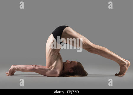 Sporty muscolare yogi giovane uomo facendo halasana, aratro pongono (yin yoga postura lumaca), studio shot su sfondo grigio scuro, laterale v Foto Stock