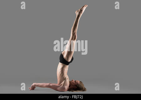 Sporty muscolare yogi giovane uomo facendo Shoulderstand non supportato pongono, Niralamba Sarvangasana, studio shot su sfondo grigio scuro Foto Stock