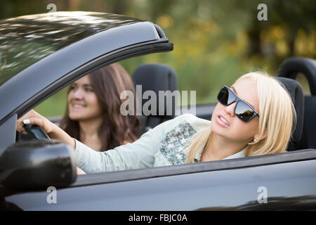 Bella ragazza bionda in occhiali da sole equitazione moderna autovettura cabriolet sul giorno di estate con passeggero sorridente bruna giovane donna, lookin Foto Stock