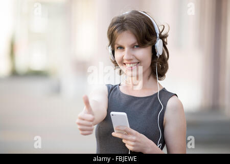 Ritratto di donna felice in bianco auricolari tenendo il cellulare in mani su estate street, ascoltando musica, friendly sorridente Foto Stock