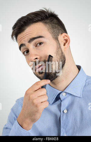 Close up ritratto di Giovane uomo barbuto la pettinatura della sua barba guardando la fotocamera. Dissaturato ritratto su grigio di sfondo per studio Foto Stock