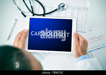 Primo piano di un medico uomo seduto alla sua scrivania in ufficio tenendo un tablet PC con la parola cardiologo nella relativa schermata Foto Stock