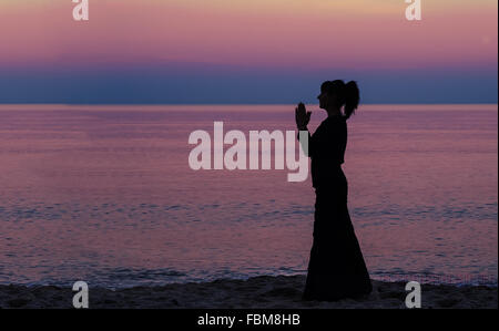 Silhouette di una donna in piedi sulla spiaggia con le mani in posizione di preghiera Foto Stock