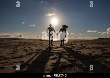 Silhouette di una giovane camminando lungo la spiaggia tenendo le mani Foto Stock