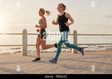 Due giovani donne che corre lungo una passeggiata sul lungomare. Montare guide giovani lavorando insieme su una strada dal mare. Foto Stock